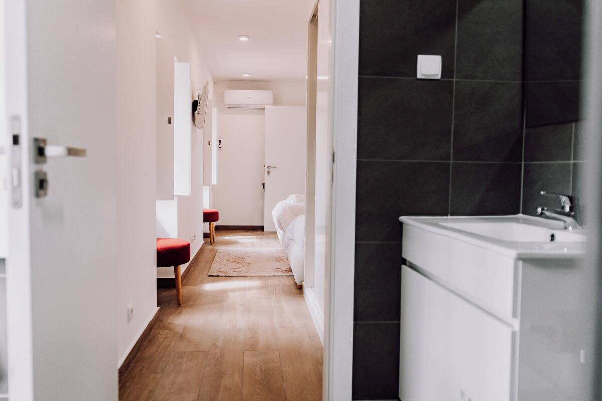 A hotel corridor with a wooden floor, a red bench, a white bed, and a bathroom with a white sink and dark tiles.