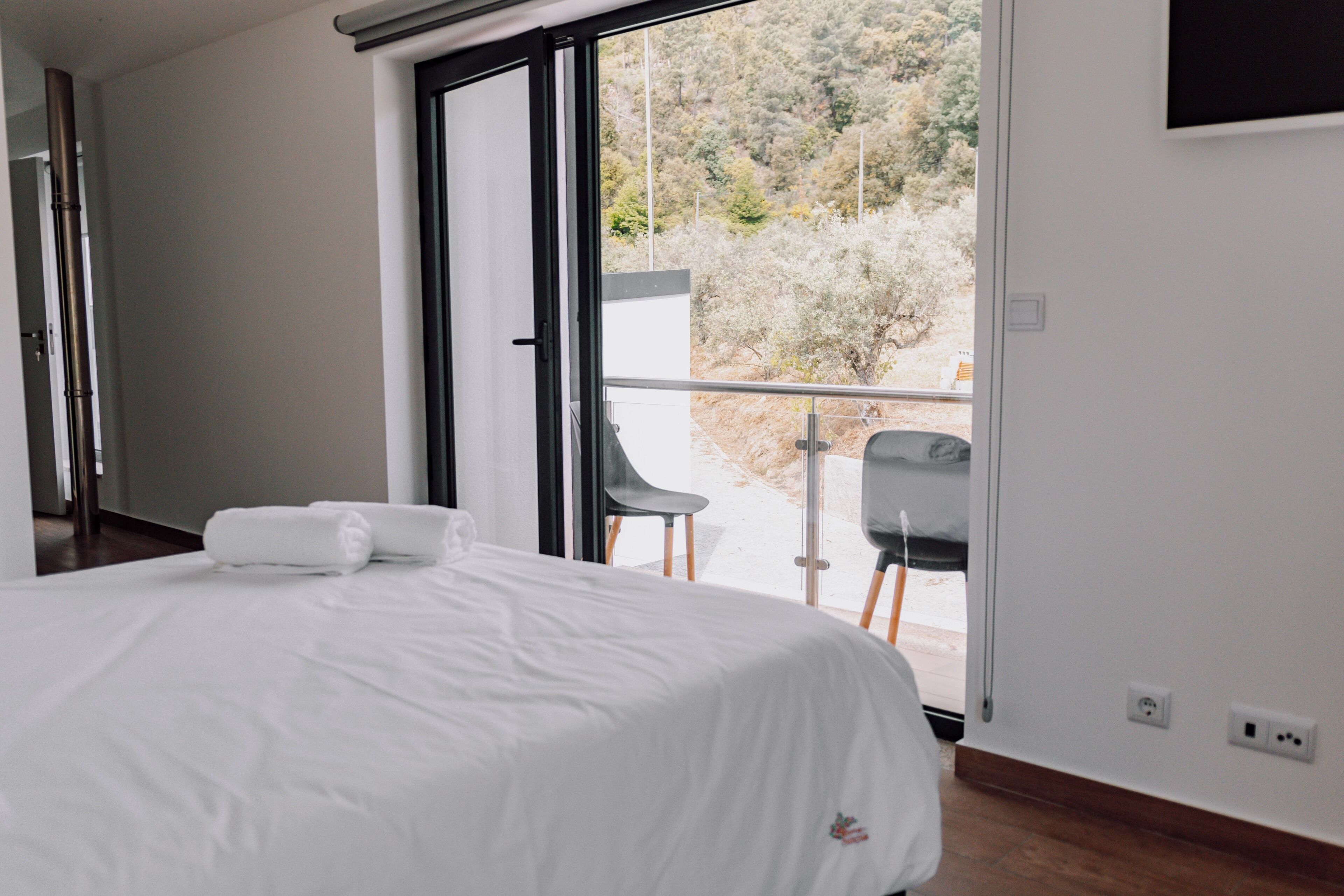 A modern hotel room with a large bed, a balcony with a table and chairs, and a view of the surrounding landscape.