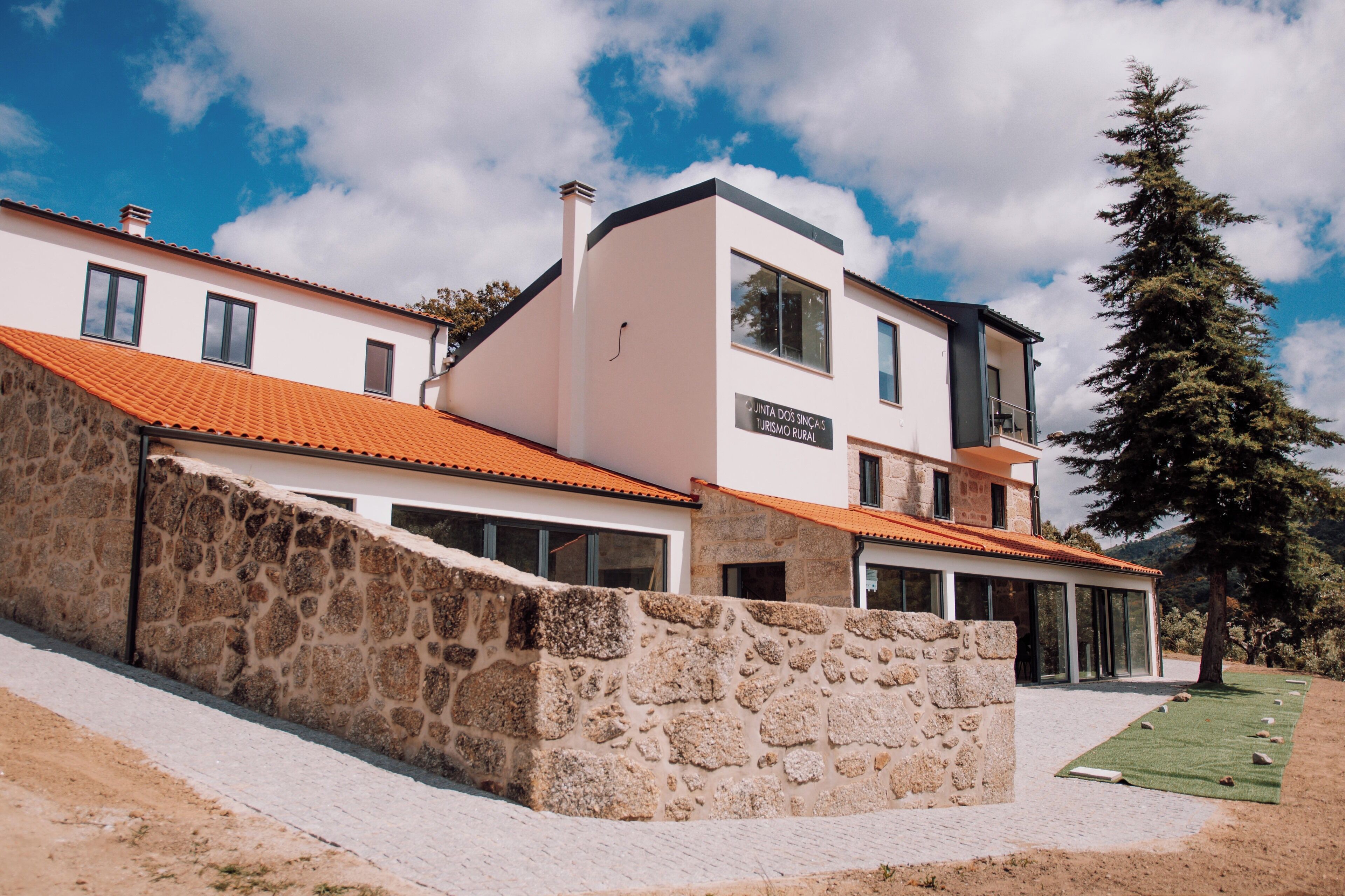 A modern building with a stone wall, a sign reading "Junta dos Ingleses Turismo Rural," and a clear blue sky.