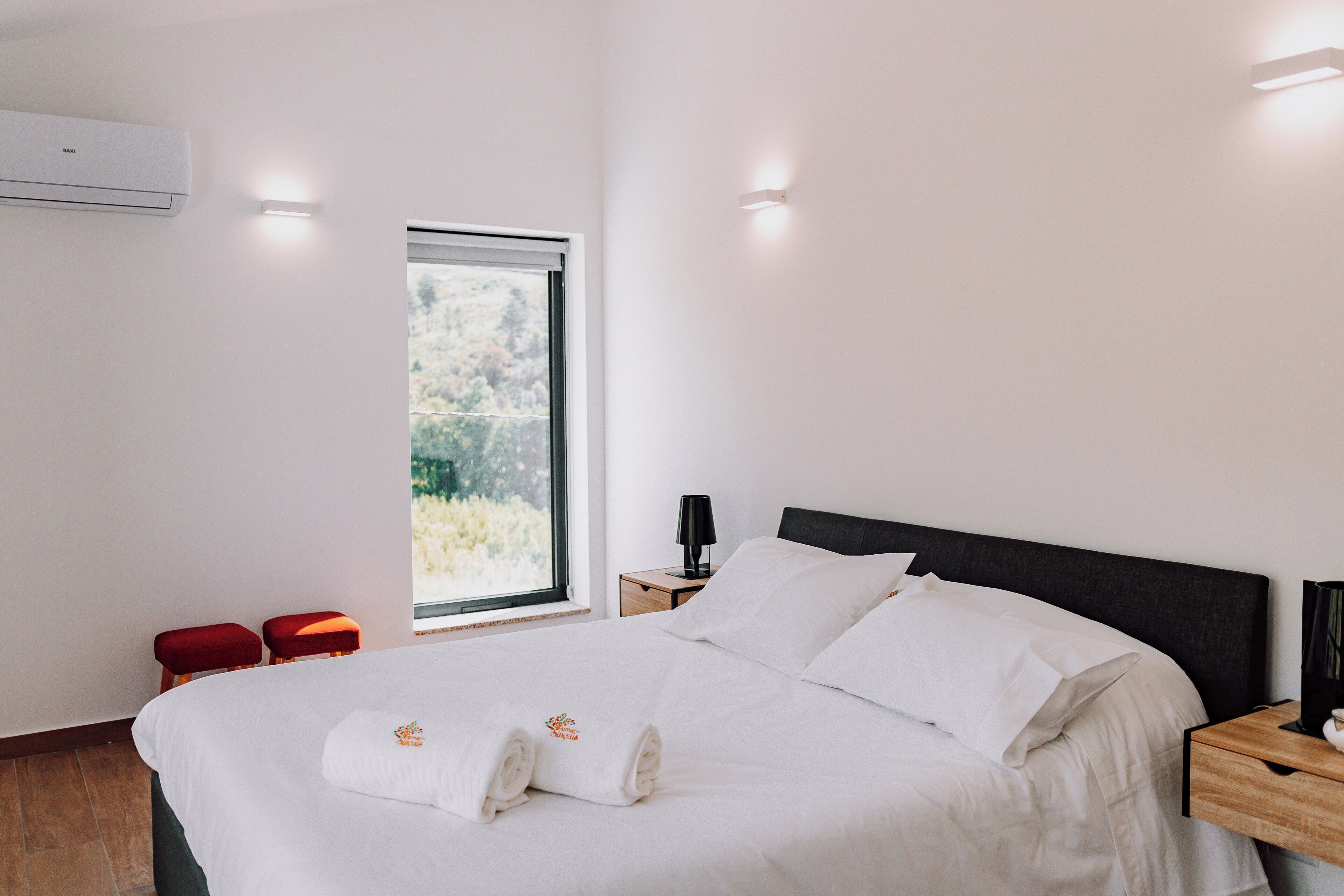 A modern bedroom with a large bed, a window with a view of greenery, a bedside table with a lamp, and a wall-mounted air conditioner.
