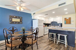 A modern kitchen with white cabinets, a blue accent wall, a dining area with a glass table, and a ceiling fan. - 1410 Waters Edge Resort 1 Bedroom Condo by RedAwning (Murrells Inlet)