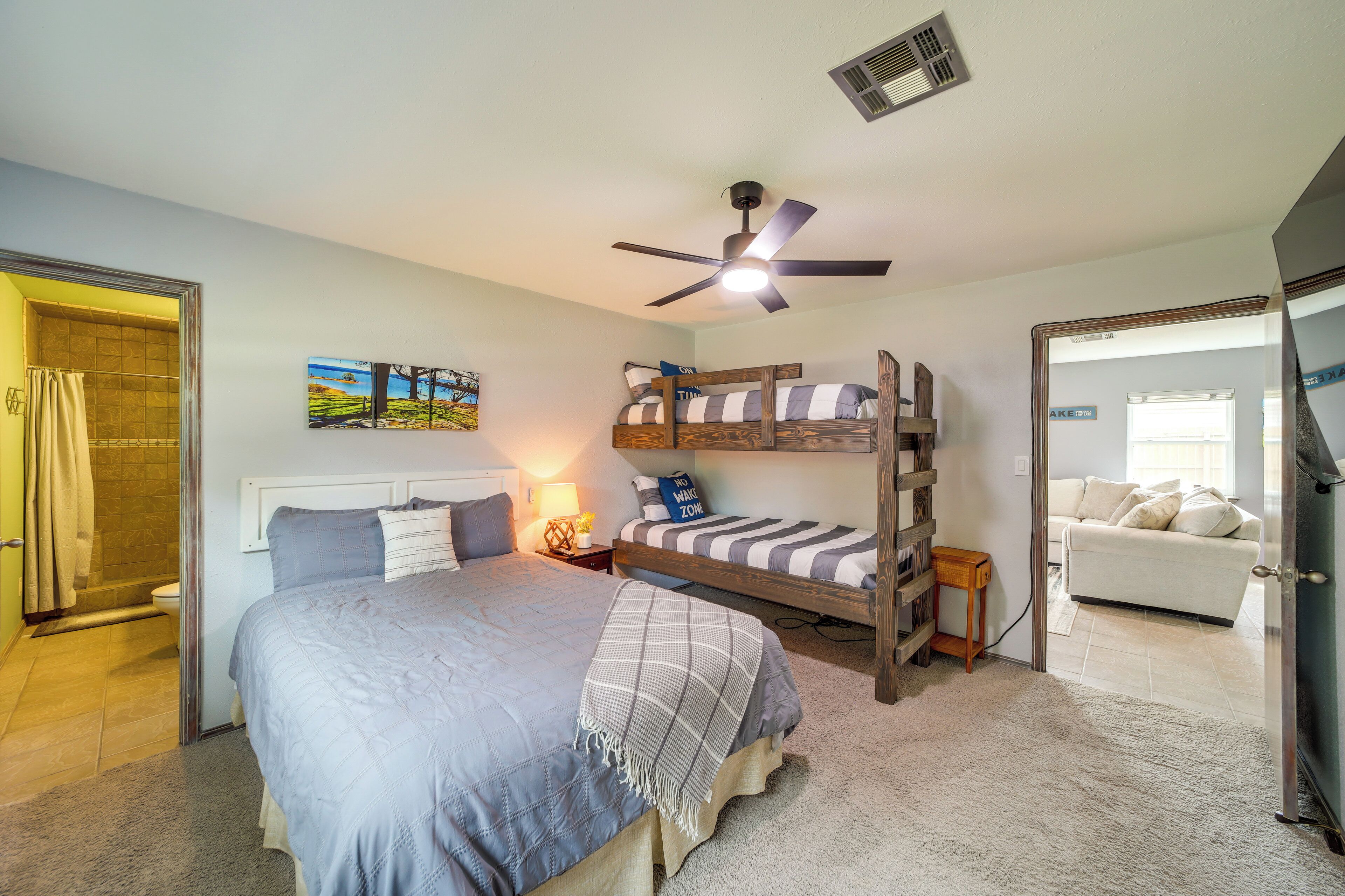 A bedroom with a bunk bed, a single bed, a ceiling fan, a bedside table with a lamp, and a view of a living area with a couch.