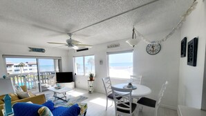 Una habitación luminosa con ventilador de techo, televisor, mesa de comedor con sillas, sofá con funda azul y vistas a la playa a través de amplias ventanas.