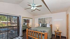 A bedroom with a wooden bed, a ceiling fan, a window with blinds, and a view of the outdoors. - Sunrise Pines Cabin | Spa · Near Village (Big Bear Lake)