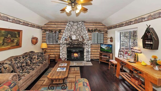 A cozy living room with a stone fireplace, wooden furniture, and a ceiling fan. - Sunrise Pines Cabin | Spa · Near Village (Big Bear Lake)