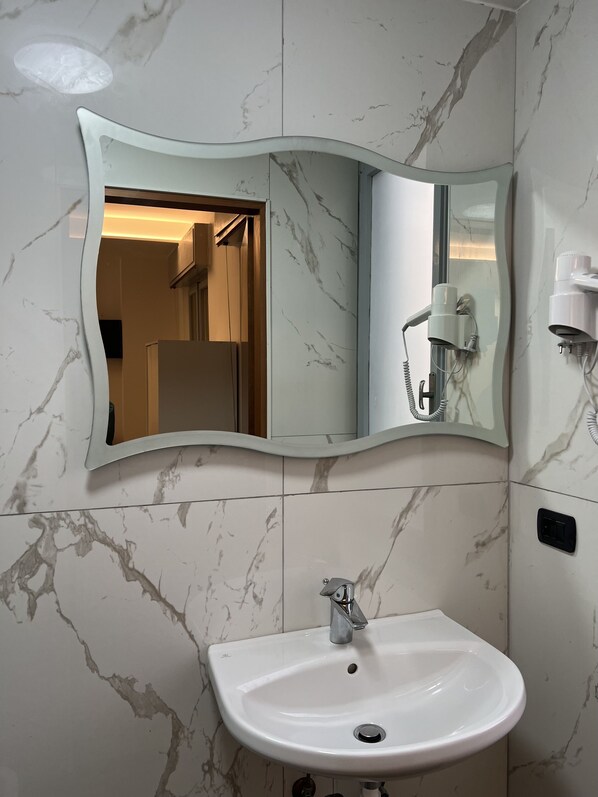 A bathroom with a marble sink, a curved mirror, and a hairdryer mounted on the wall. - Hotel Mirò (Rimini)