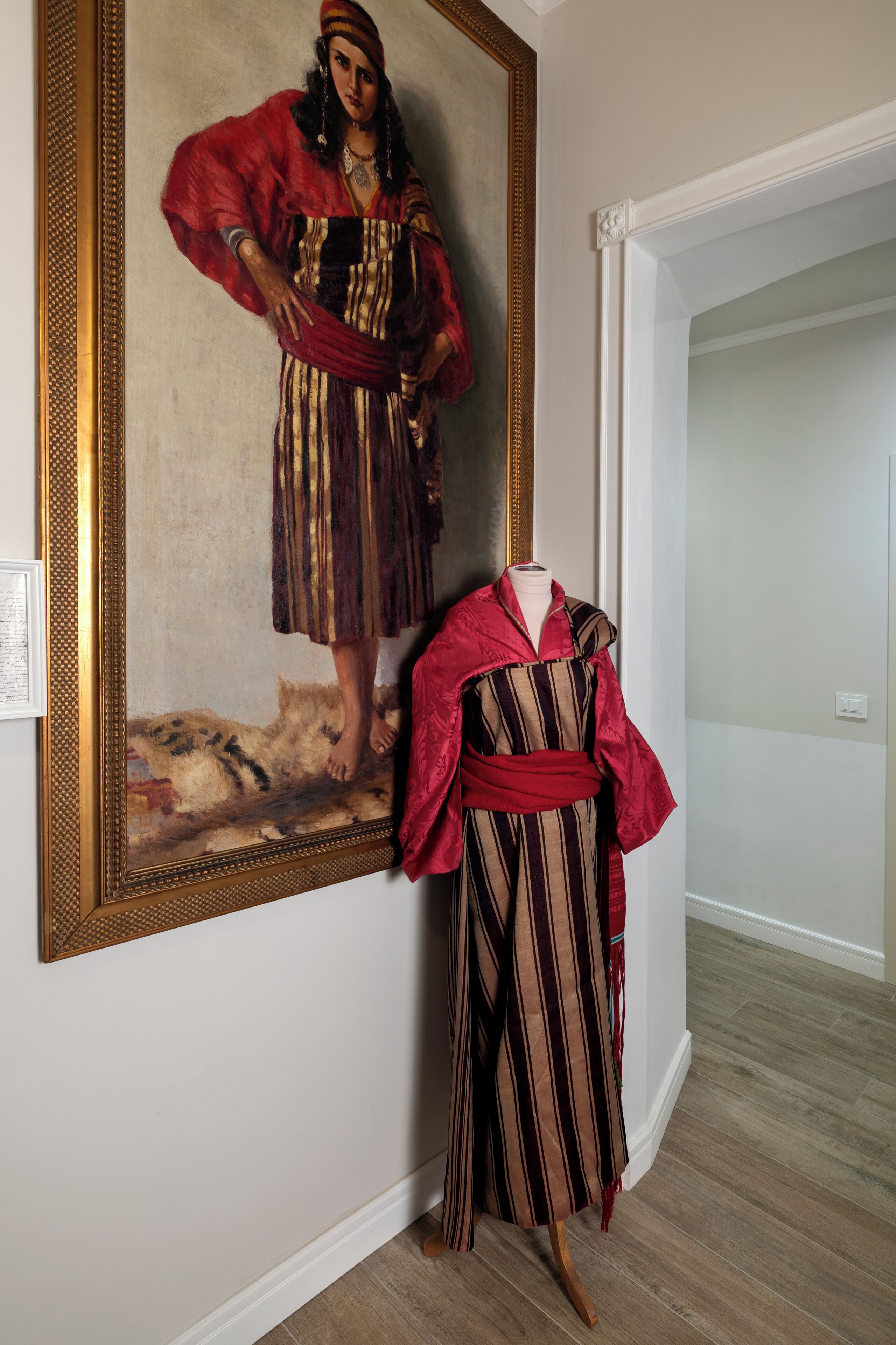A room with a large framed painting on the wall, a mannequin dressed in a striped garment, and a wooden floor.