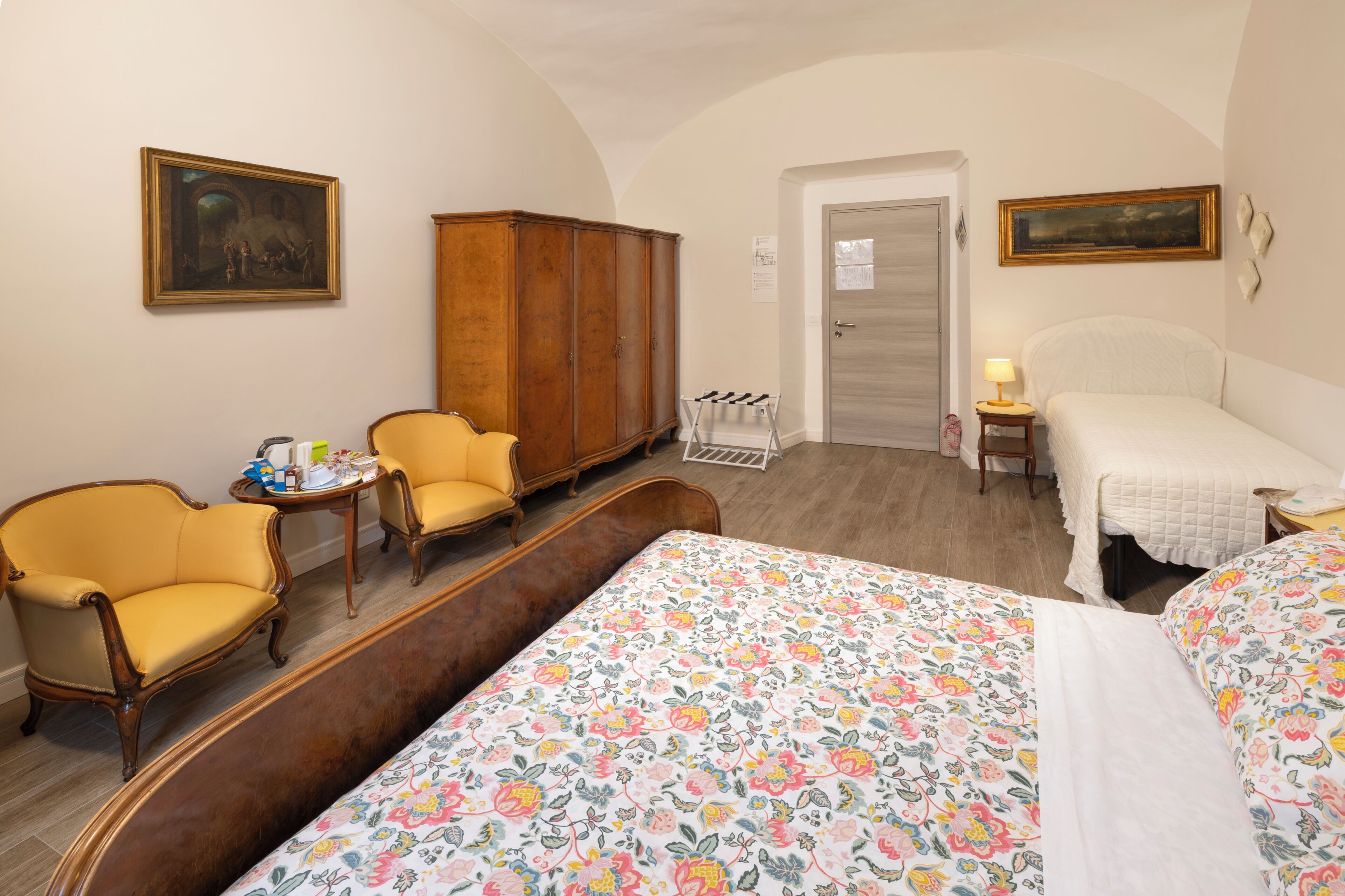A hotel room with a bed, two armchairs, a wooden wardrobe, a small table with a lamp, and a door leading to another room.