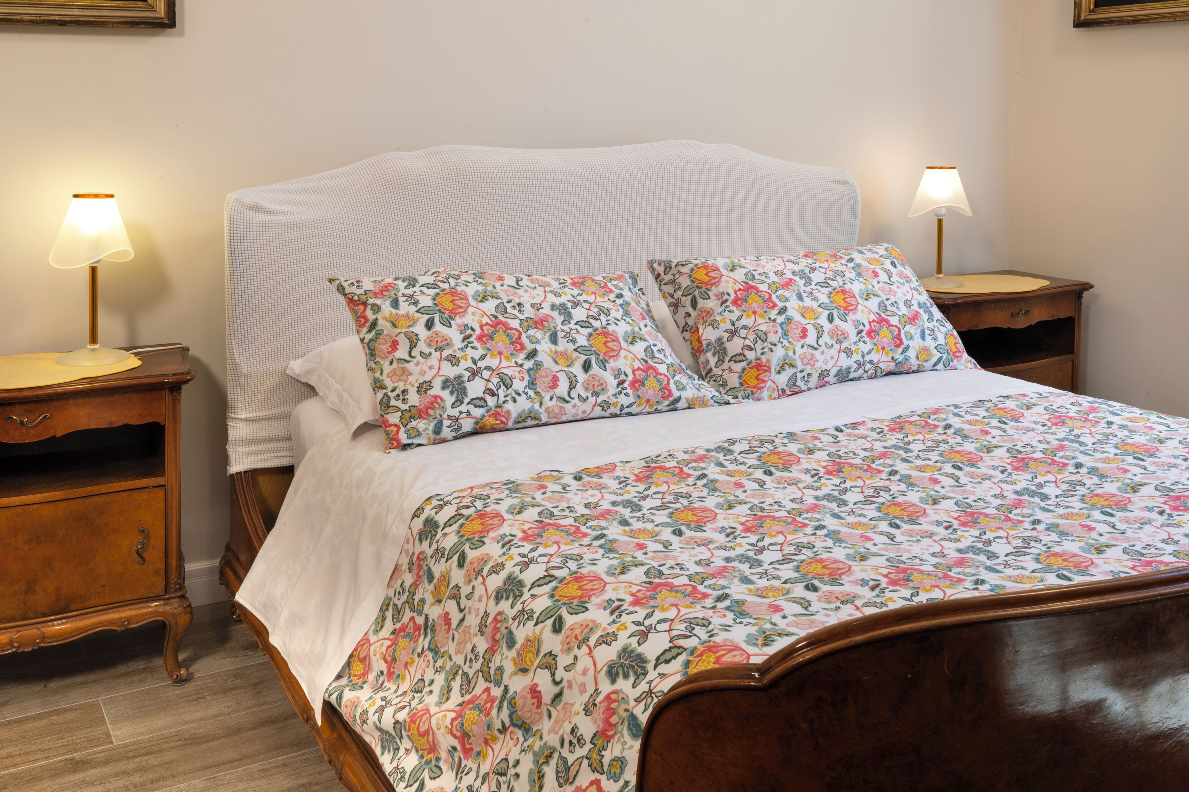 Una cama bien hecha con ropa de cama floral, dos mesitas de noche con lámparas y un pequeño mueble de noche de madera con un cajón.