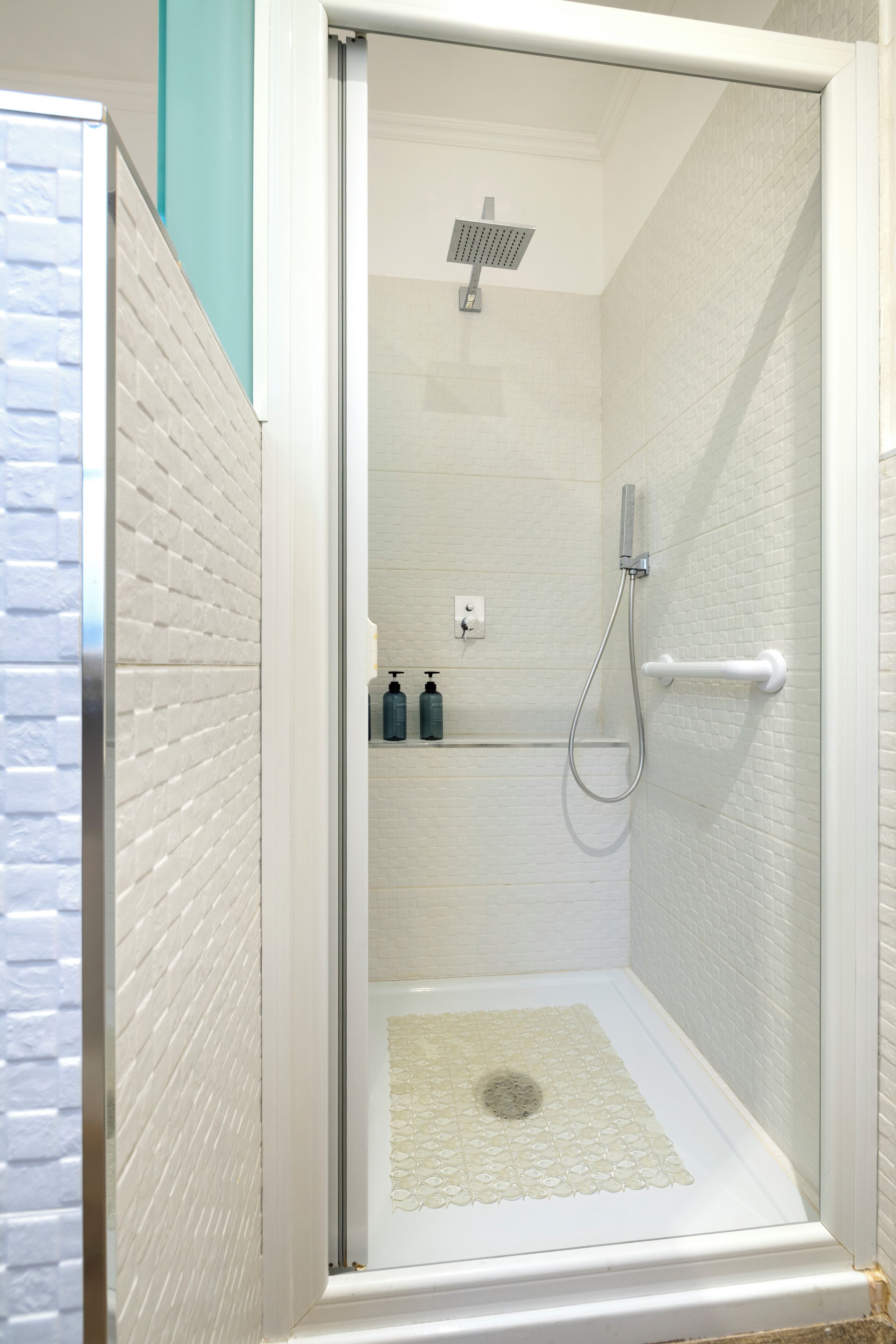 Ducha a pie con puerta de vidrio, paredes de azulejos blancos y un estante pequeño con artículos de tocador.