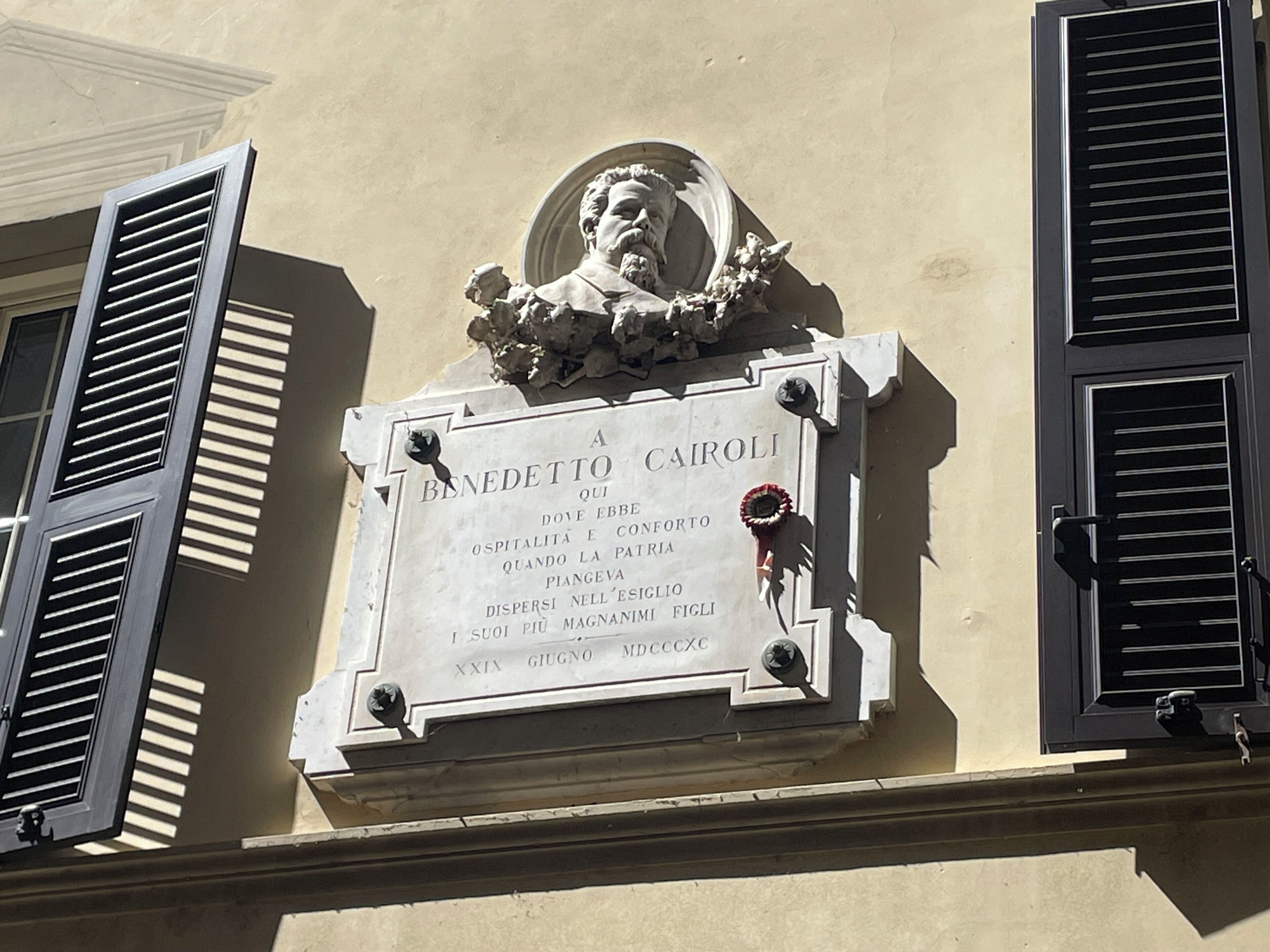 A plaque with a bust and inscription, mounted on a building facade with shutters.