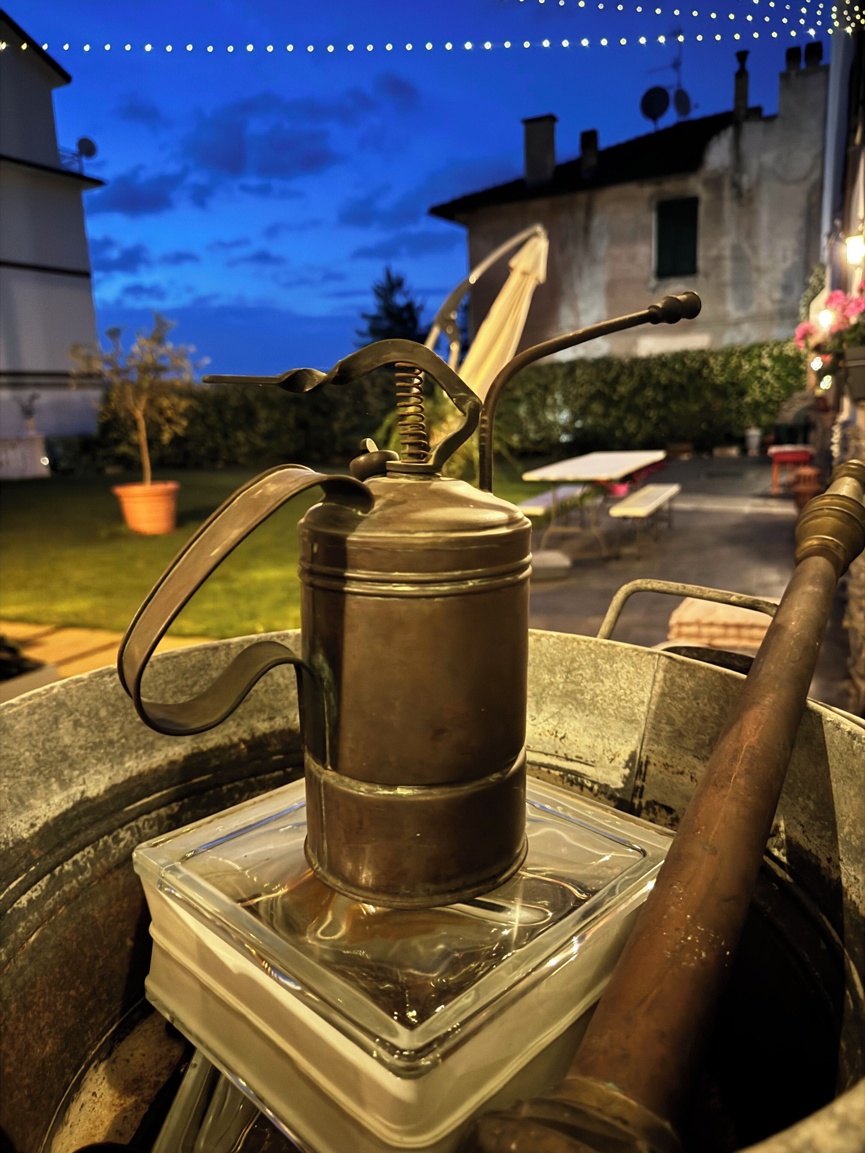 A metal coffee pot on a clear plastic container, set outdoors with a house and greenery in the background.