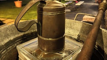 Une cafetière en métal posée sur un récipient transparent en plastique, le tout installé à l’extérieur, avec une maison et de la verdure en arrière-plan.