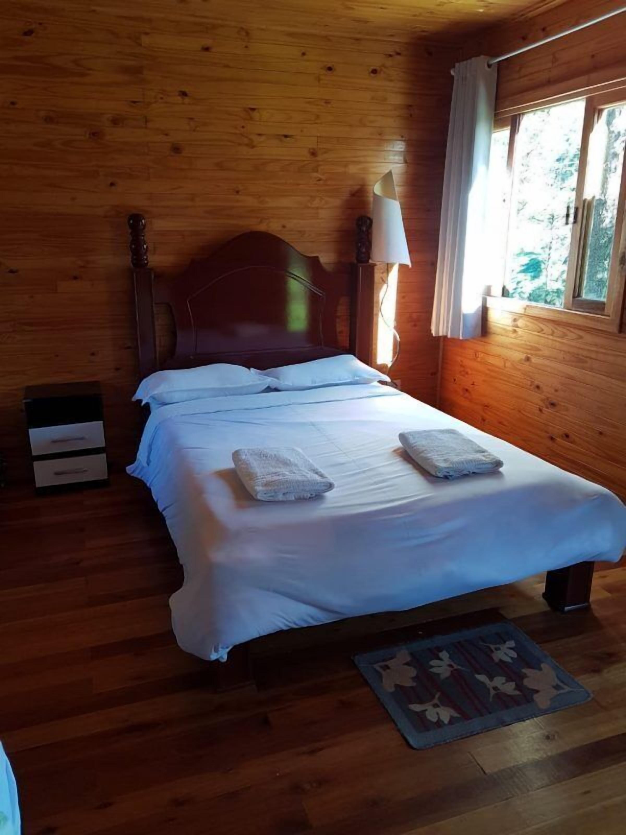 Quarto com painéis de madeira, uma cama, uma mesa de cabeceira, um abajur e uma janela com cortinas.