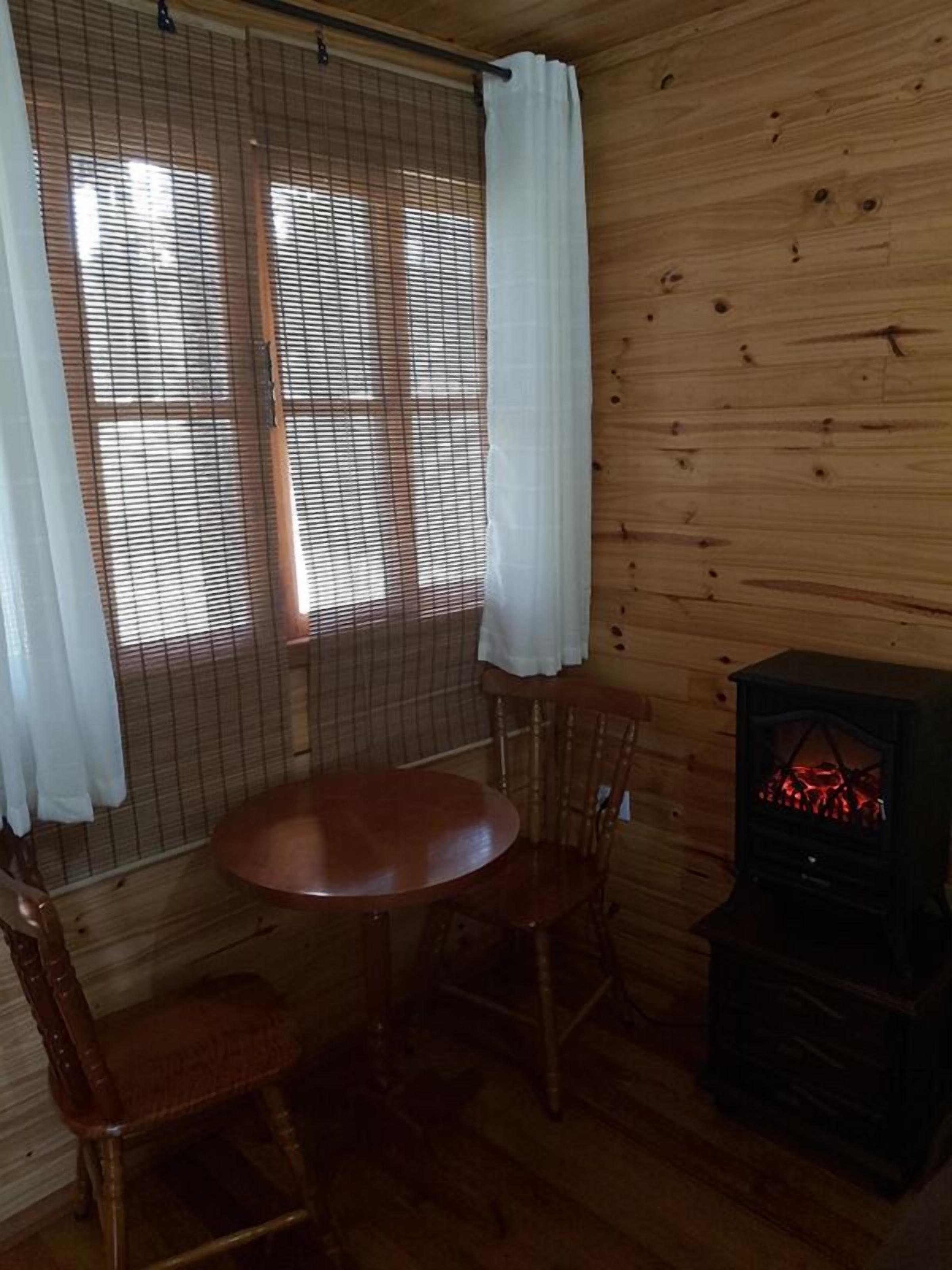 L’intérieur d’un chalet en bois, avec une table ronde et deux chaises, un poêle à bois et une fenêtre équipée de stores et d’un rideau blanc.