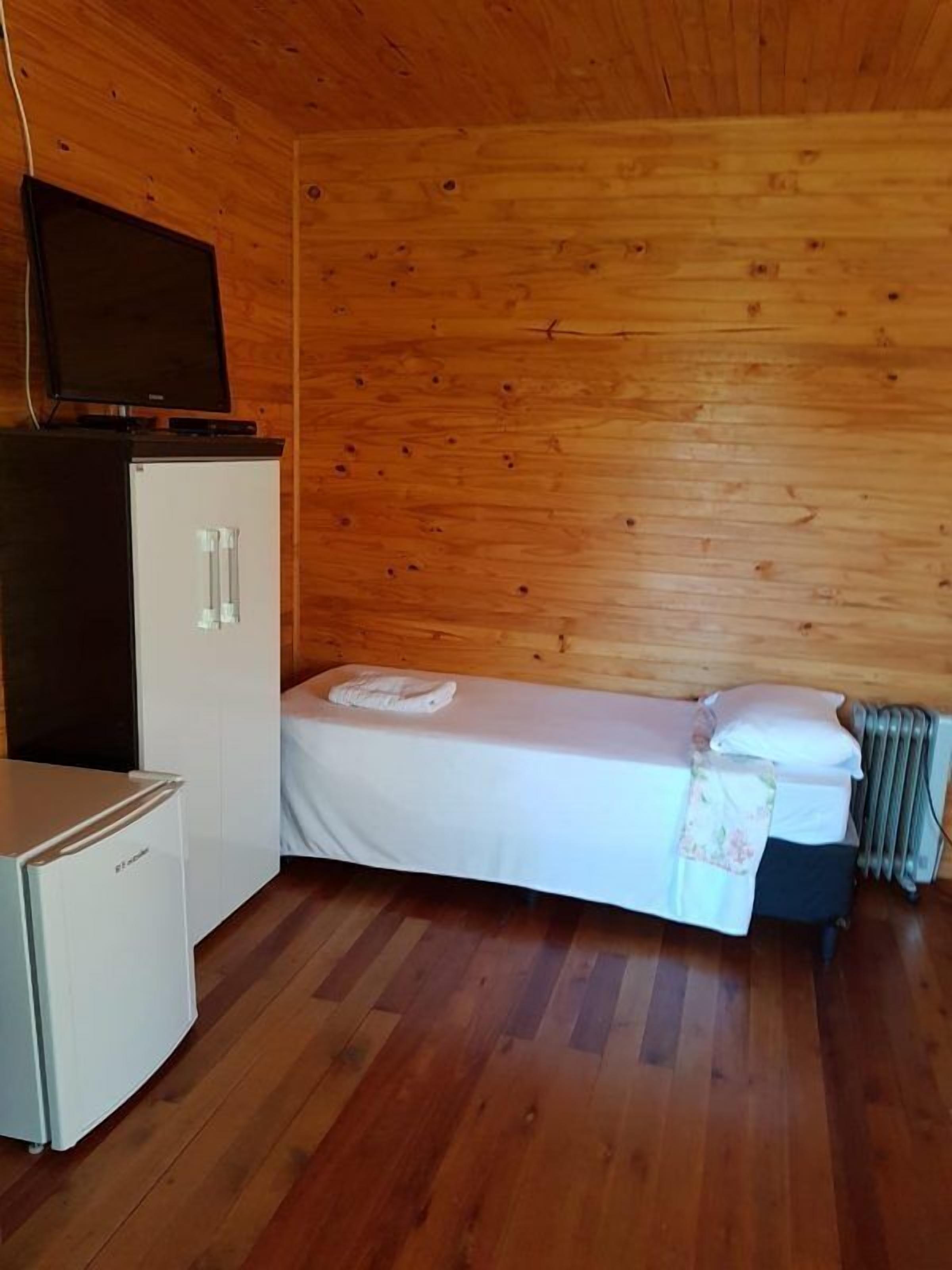 Una habitación pequeña y compacta con decoración de madera, una cama individual con ropa blanca, un refrigerador blanco pequeño y un televisor sobre un mueble.