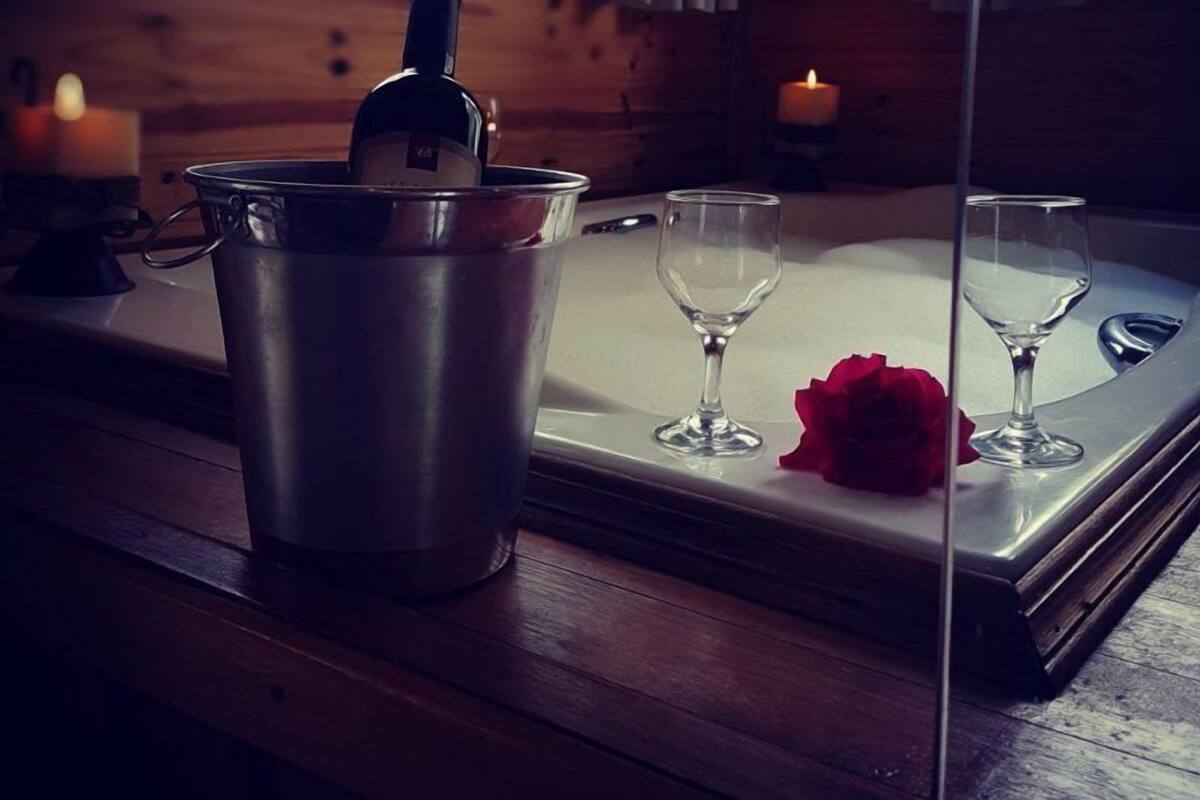 Um quarto acolhedor com decoração em madeira, uma janela com cortinas, uma vela, uma garrafa de vinho em um balde e duas taças de vinho sobre uma bandeja com uma rosa vermelha.