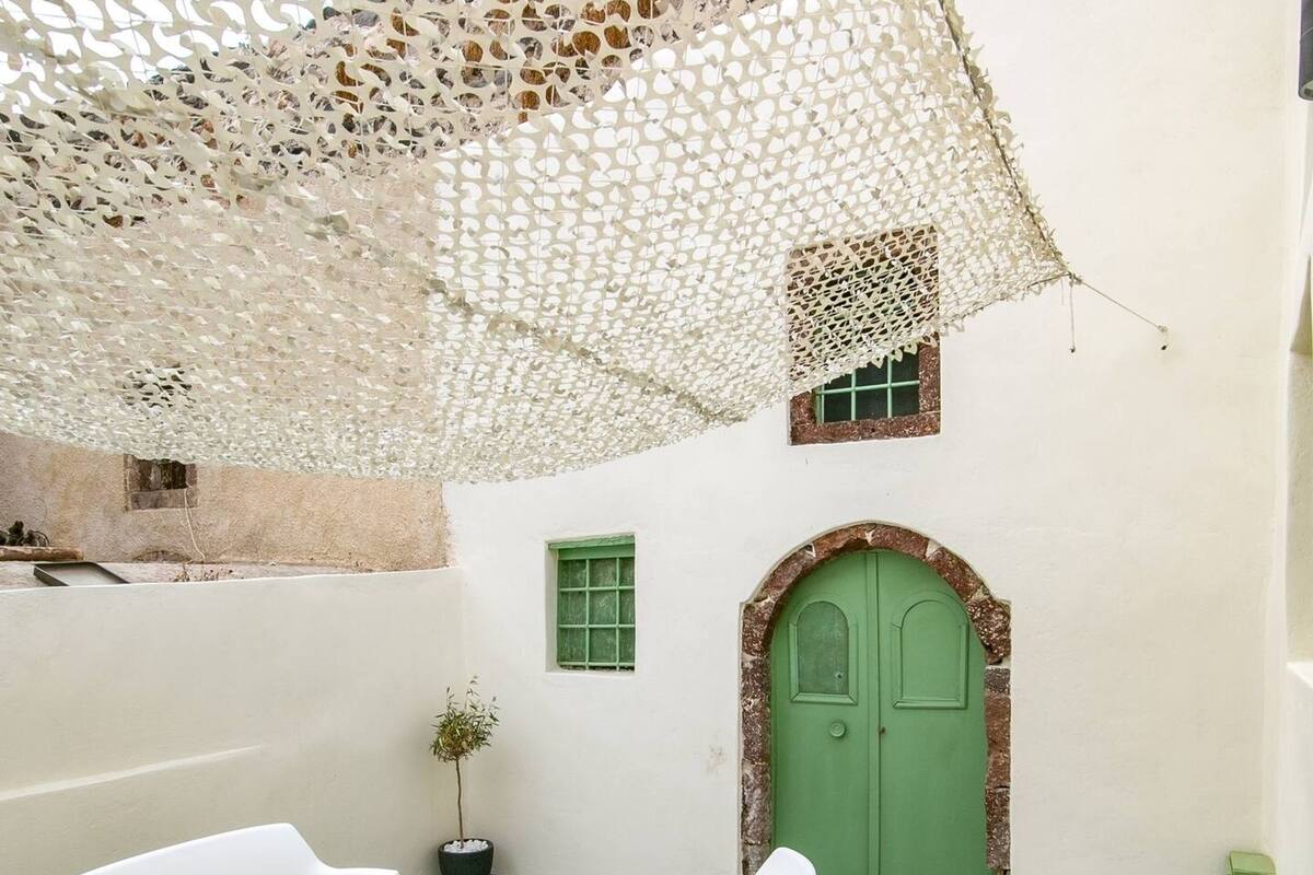 A courtyard with a white table, two white chairs, a green door, and a white wall with a small window.