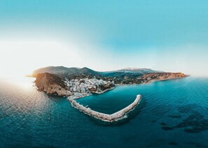 Vista aérea de un pueblo costero con un puerto en forma de corazón, rodeado de montañas y aguas azul cristalina.