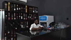 Un coin bar où un homme est assis au comptoir, entouré d’étagères remplies de bouteilles d’alcool.