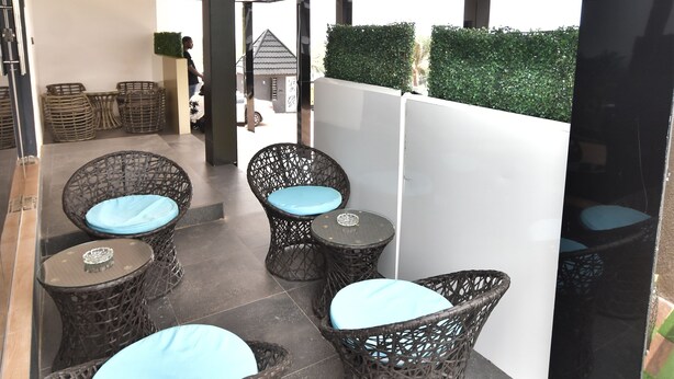 Un espace extérieur moderne, aménagé avec des fauteuils en osier et une table en verre, entouré d’un paravent blanc et d’une haie.