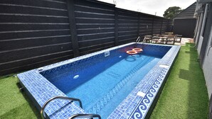 Une piscine rectangulaire aux carreaux bleus, dotée d’un escalier, entourée de gazon synthétique et d’une clôture noire.