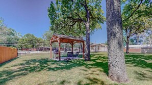 A wooden gazebo with benches, surrounded by a fenced yard and trees.