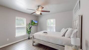 A bedroom with a bed, a ceiling fan, two windows with blinds, a plant, and a wall-mounted TV.