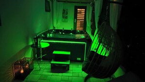 Ein schwach beleuchteter Raum mit grünem indirektem Licht, einem Bett, einem Hängesessel und einem kleinen Tisch mit einer Lampe.