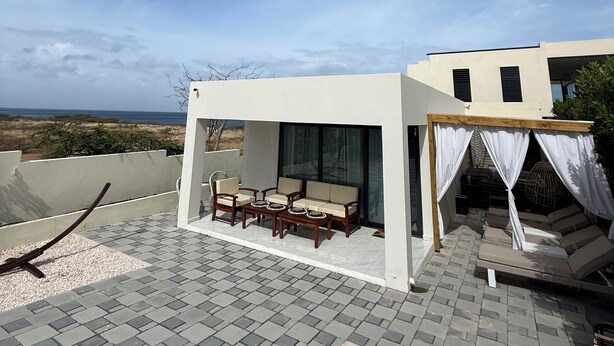 Ein moderner Außenbereich mit einer Couch und Sesseln, einer Pergola mit weißen Vorhängen und Blick auf das Meer.