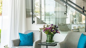 Un salon moderne avec deux fauteuils gris, une table basse blanche et un vase rempli de fleurs. On aperçoit un escalier avec des rampes en verre à l’arrière-plan.