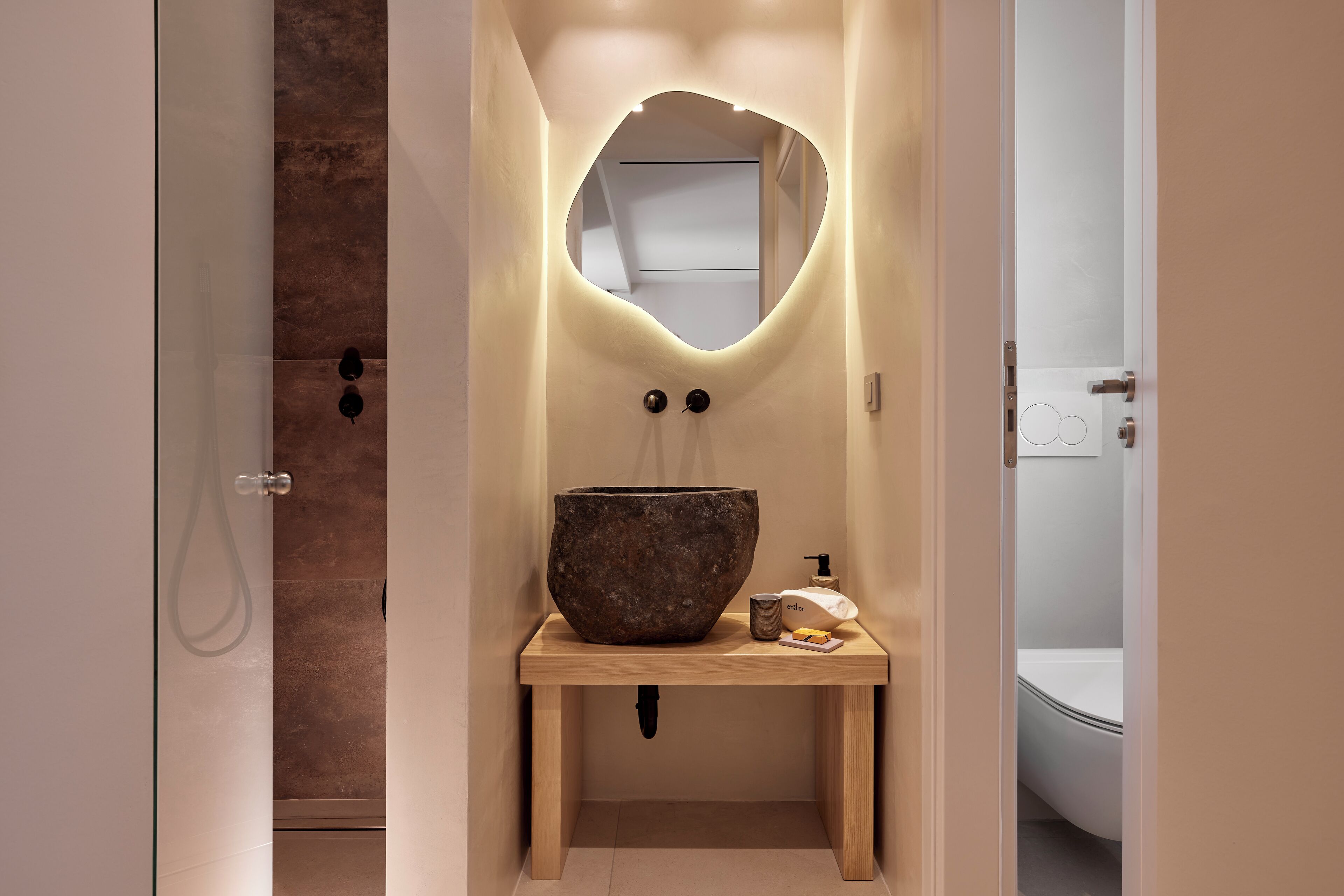 Ein Badezimmer mit einem großen, rustikalen Steinsink, einem Holzwaschtisch und einem Spiegel mit ungewöhnlicher, organischer Form.