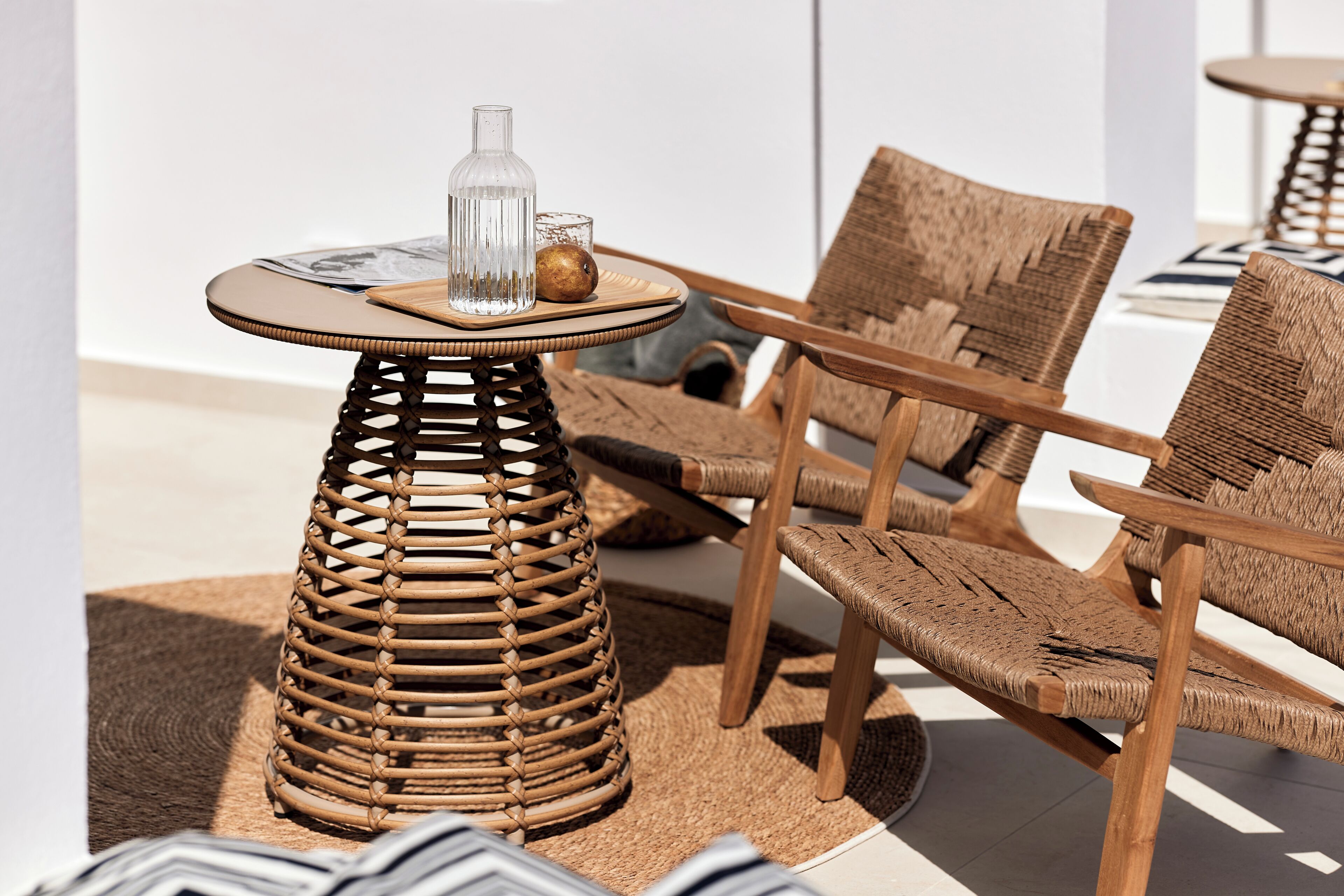A wicker chair and table set on a woven rug, with a glass bottle and a bowl on the table.