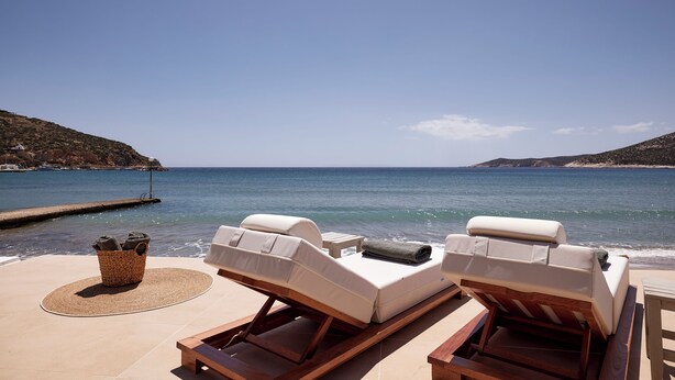 Area relax in spiaggia con due sdraio, un cesto di vimini con asciugamani e vista sul mare e le colline.