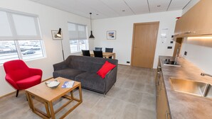 Una sala de estar moderna con un sofá, una silla roja y una mesa de madera. Se observa una zona de cocina con un fregadero y armarios.