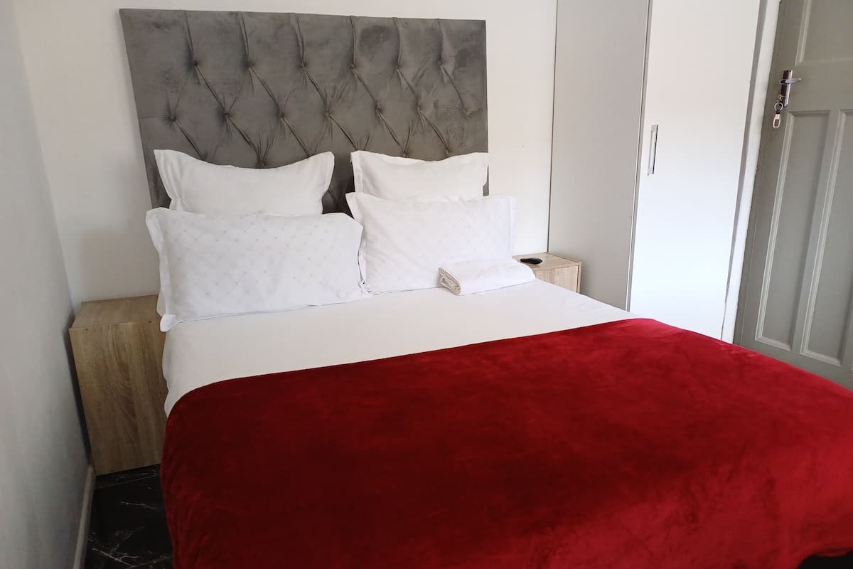 Een bed met een grijs gewatteerd hoofdbord, witte kussens en een rode deken.