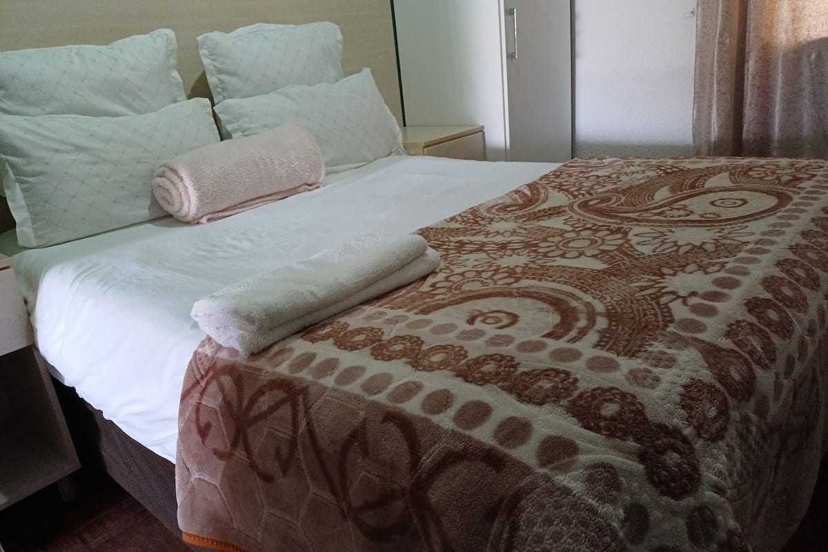 Een netjes opgemaakt bed met een beddekleed met patroon en kussens, een nachtkastje en een garderobe.