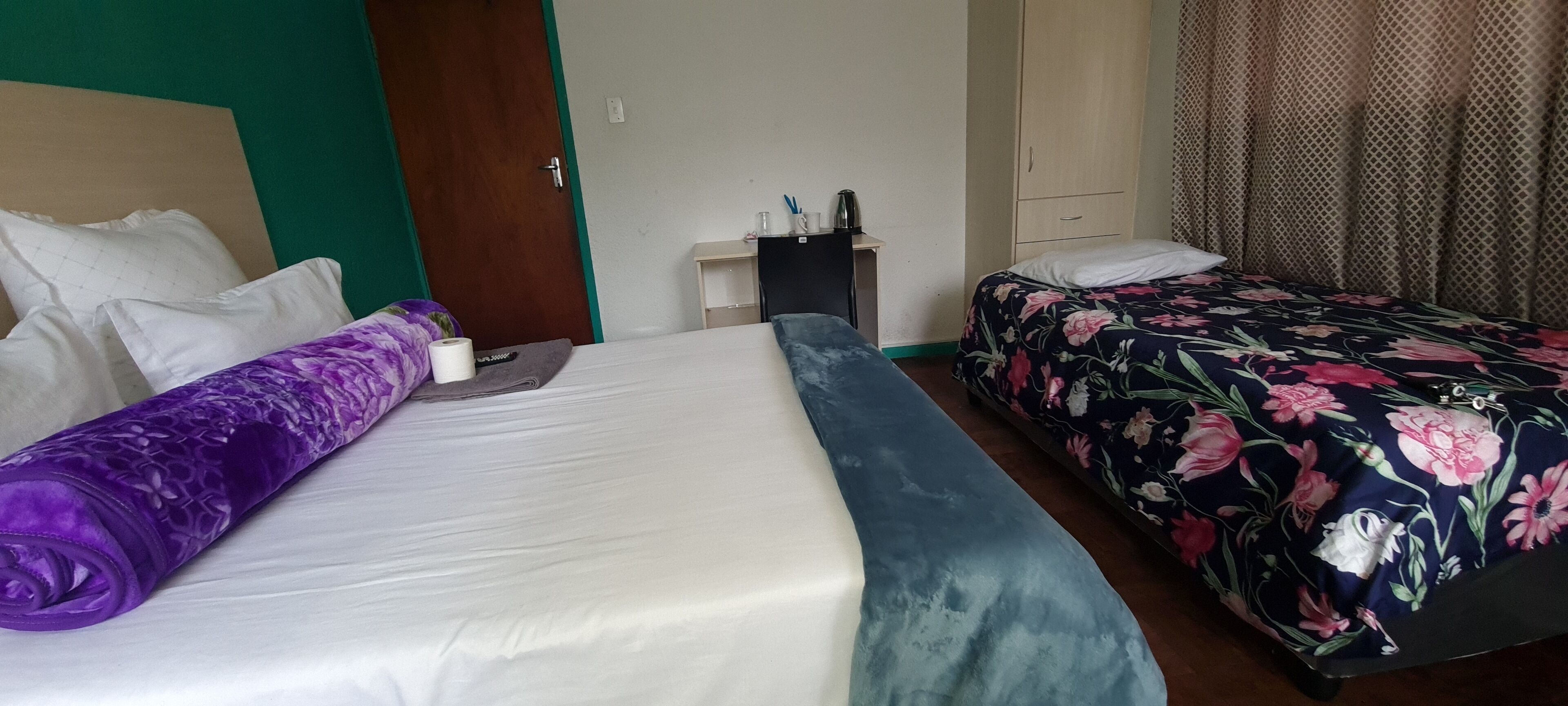 Habitación de hotel con dos camas, una con una manta morada y la otra con un edredón floral. Hay una mesita con una tetera y tazas, y un armario al fondo.
