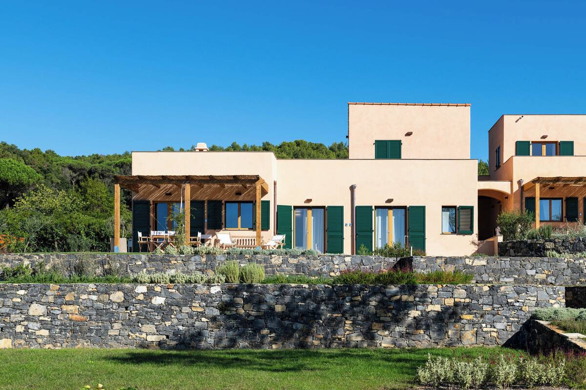 A modern building with a stone wall in front, multiple doors with green shutters, and a clear blue sky.