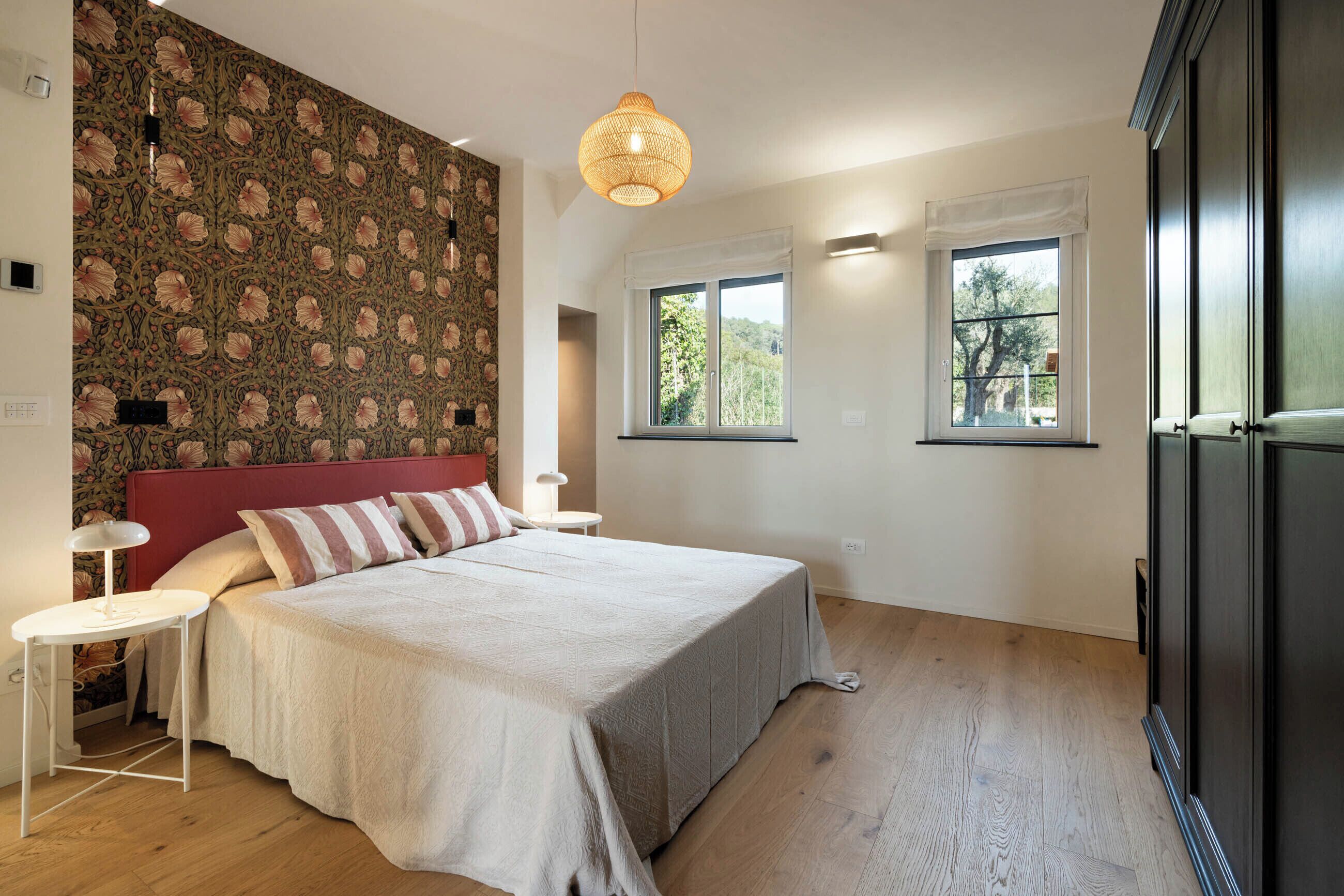 A bedroom with a large bed, two bedside tables, a red headboard, a patterned wallpaper, and two windows with curtains.