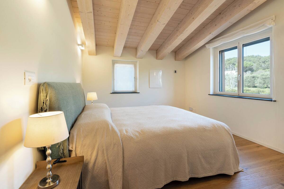 A bedroom with a bed, a nightstand with a lamp, a window with a view of greenery, and wooden beams on the ceiling.