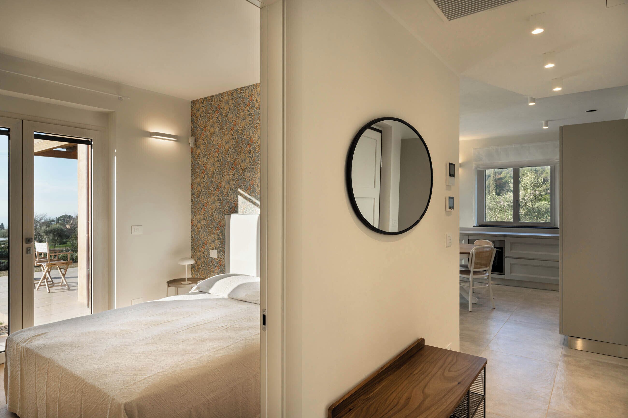 Une chambre d’hôtel moderne avec un grand lit, un miroir rond et une vue sur l’extérieur grâce à une porte-fenêtre coulissante.