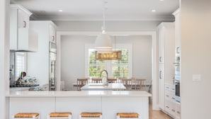 Une cuisine moderne avec des meubles blancs, un îlot central doté de tabourets, un luminaire suspendu et une vue sur la verdure à l’extérieur.