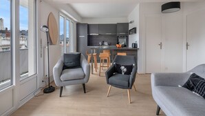 Un salon moderne avec des canapés et des fauteuils gris, un coin repas avec des chaises en bois, et une cuisine avec des meubles sombres et un plan de travail blanc.