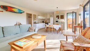 A spacious living area with a sofa, coffee table, and dining table. A surfboard is mounted on the wall, and the kitchen is visible in the background. - Saint-Vincent-sur-Jard, house for 5 (Saint-Vincent-sur-Jard)