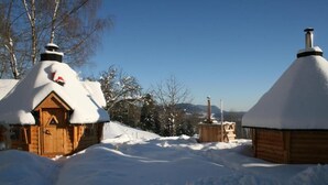 Een houten hut met een met sneeuw bedekte dak en schoorsteen, omringd door besneeuwde grond en bomen.