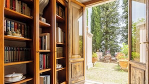 Eine Bibliothek mit offenen Regalen, gefüllt mit Büchern, und Blick auf einen Garten durch eine offene Tür.