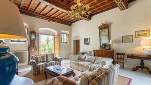 Ein geräumiges Wohnzimmer mit gestreiften Sofas, einem Couchtisch und einem Kronleuchter. Ein großes Spiegel, eine Uhr und ein Fenster mit Blick auf Grün sind ebenfalls vorhanden.
