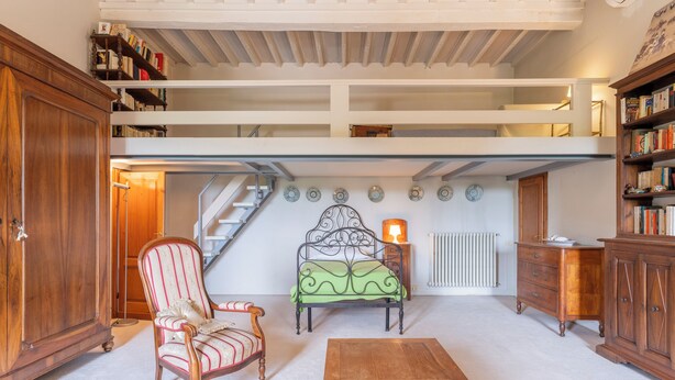 A spacious loft with a high ceiling, a staircase, a green sofa, a wooden chair, a coffee table, and a bookshelf.