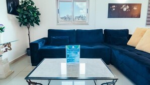 Un salon avec un canapé bleu foncé, une table basse en verre, une plante en pot et une vue sur la ville par la fenêtre.
